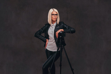 Smart blonde female photographer in trendy clothes posing while leaning on a professional camera with a tripod at a studio, looks at the camera.