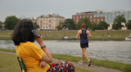 Czarnowłosa kobieta w żółtekj koszulce i kolorowych cienkich spodniach siedzi, bokiem, na nabrzeżu rzeki w mieście, trzyma okulary słoneczne, patrzy za uprawiającą jogging młodą kobietą w szortach - obrazy, fototapety, plakaty
