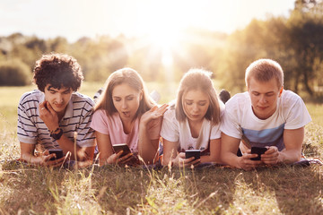 Addicted teenagers being always in touch, lie on green grass, use mobile phones during rest outdoor, have different facial expressions, surf internet or type text messages, focused in their devices