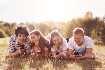 Joyful friends have fun together, use modern cell phones, watch funny videos, being addicted from modern technologies, have good relationship, enjoy hot summer day and nice nature in countryside