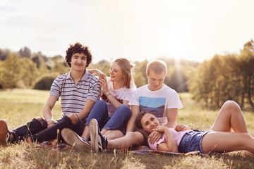 Outdoor shot of cheerful youth couples enjoy togetherness and spare time during summer day or weekend, sit closely to each other, admire beautiful nature, have pleasant talk. Teenagers and relations