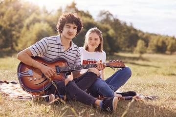 Youth, teenagers and entertainement concept. Handsome male plays guitar and sings songs to his girlfriend, look happily at camera, have joyful expressions, enjoy togetherness and beautiful nature