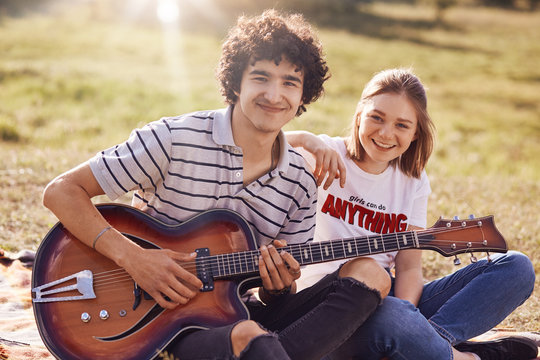 Two happy friends have joyful expression, gentle smiles on faces, recreat during summer time outdoor, play guitar and sing popular songs, can`t imagine their lives without music. Rest concept