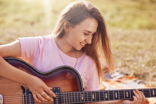 Horizontal shot of happy lovely female with positive smile plays guitar, learns new song, recreats oudoor in meadow, dressed in casual t shirt, has joyful expression. People, music, rest concept