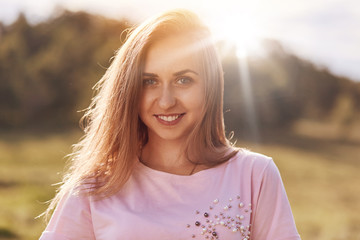 Close up shot of happy female with charming smile has attractive look, blue eys and white teeth, dressed in casual clothes, stands against sunlight, expresses positive emotions and feelings.