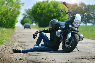 Biker in outfit sits next to his motorcycle on the road. Motorcyclist rests near his motorbike.