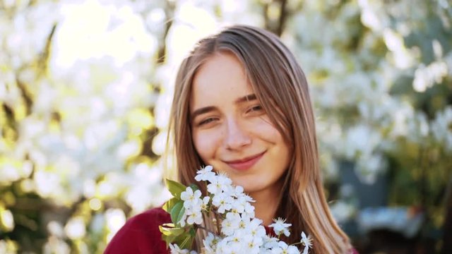 Cute Ukrainian young woman with blue eyes holding twigs with white flowers, looking at camera and smiling. Likable girl in the garden. Feminine image.