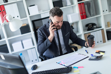 Naklejka premium A man is sitting at the desk at the office, talking on the phone and holding a glass of coffee in his hand.