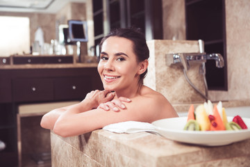 Happy young female lying in bath with folded hands. She is leaning on towel with smile and having fruit plate beside her 