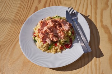A tasty and healthy coucous salad consist with cucumber, choped tomatoes and red pepper and poached salmon pieces served on the white plate and stainless steel cutlery. 