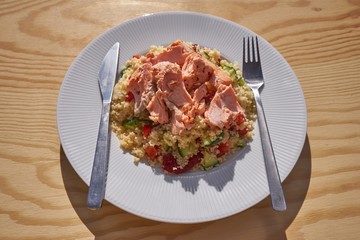 A tasty and healthy coucous salad consist with cucumber, choped tomatoes and red pepper and poached salmon pieces served on the white plate and stainless steel cutlery. 