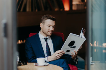 Handsome young businessman looks attentively at magazine, dressed in formal suit, drinks aromatic...