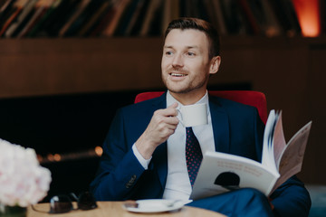 Horizontal shot of happy businessman reads popular magazine in cafeteria, drinks aromatic coffee, dressed in formal suit, has pleasant smile, wears formal black suit, enjoys recreation time.