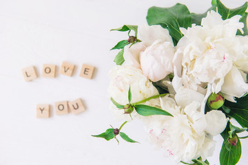Flat lay focus on fresh bouquet of white peonies and blurred Love you lettering spelled in wooden blocks on light background. Love words. Valentine's day. Top view. Copy space, selective focus.
