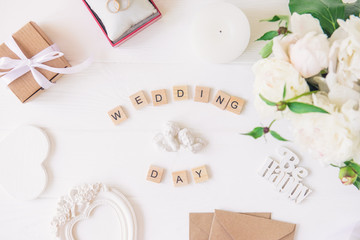 Layout with Wedding day spelled in wooden blocks, statuette of two lovely angels, giftbox, rings, postcards, heart shaped frame and white peonies on the wooden table. Invitation, greeting concept.