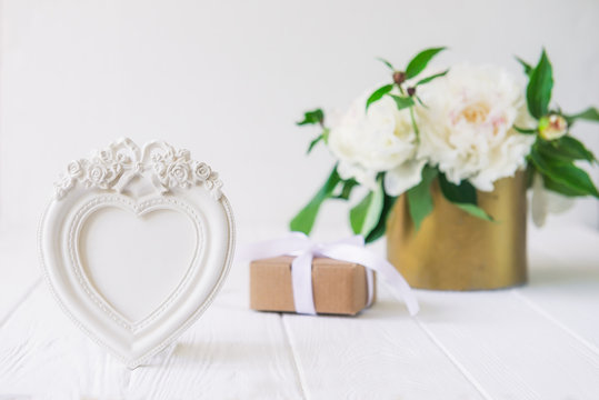 Heart shaped photoframe with plaster flowers,gift box with ribbon and vintage bowl with white peonies bouquet on the wooden table. Love background. Greeting card. Copy space, selective focus.