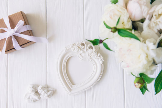 Top view vintage heart shaped photo frame, statuette of two antique lovely angels, giftbox and white peonies bouquet on the wooden table. Love background. Wedding concept. Copy space, selective focus.