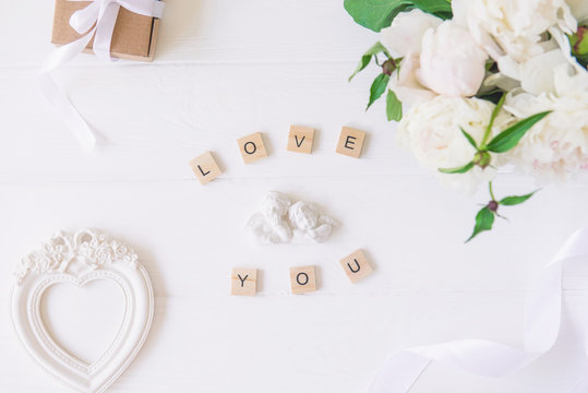 Top view Love you lettering with statuette of two little antique lovely angels, vintage heart shaped photo frame, giftbox and white peonies bouquet on the wooden table. Love background. Copy space.