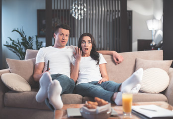 Obraz premium Full length portrait of surprised young couple is watching movie together at home. They are looking forward with shock while sitting on couch and embracing 