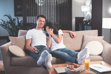 Oh my god. Terrified woman is closing her face by palm while watching interesting move together with her husband. Man is embracing wife and smiling