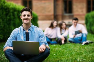 Male university student sitting on grass with laptop in front of friends