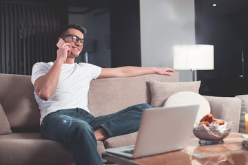Cheerful young guy is enjoying communication by smartphone. He is sitting on couch and laughing. Laptop and food is on table. Relaxation and domestic comfort concept 