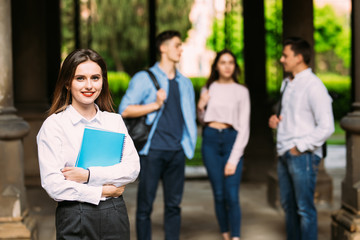 Beautiful young college student standing in front of her friends holding her books at campus....