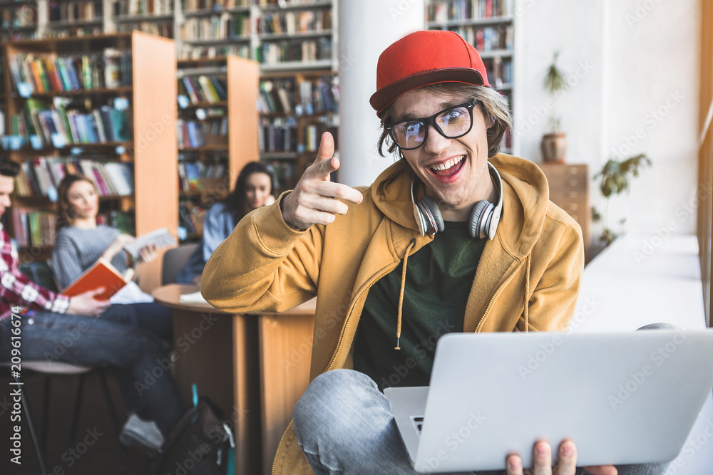 Poster Portrait of satisfied male pointing finger in camera while having job with notebook computer in library