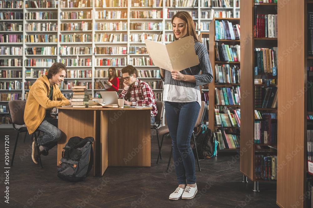 Poster outgoing woman reading information while standing in library. happy guys studying at table. they usi