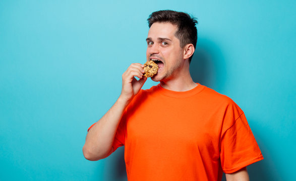 Young handsome man in orange t-shirt with cookie. Studio image on blue background