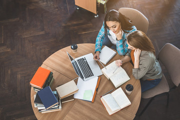 Top view happy ladies looking through volume while sitting at desk with notebook computer