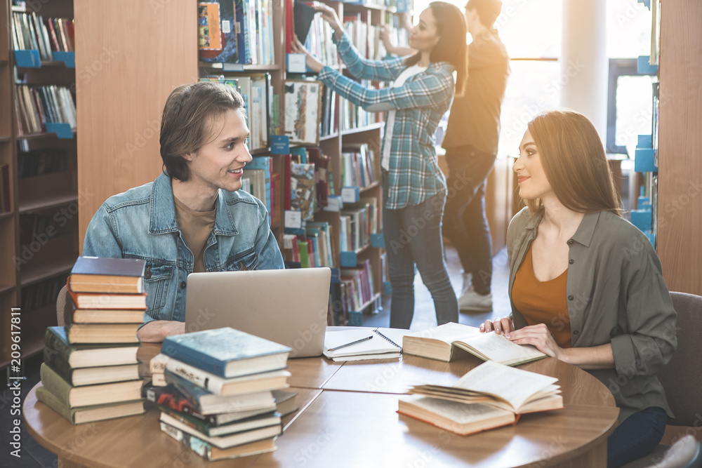 Poster side view optimistic guy speaking with smiling girl while using laptop. she reading book. they sitti
