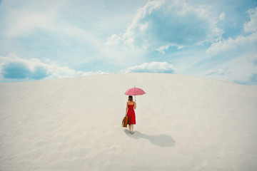 Young woman in red dress with umbrella and suitcase on the beach. Travel concept image on sand