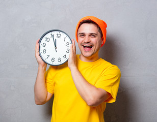 Young handsome man in yellow t-shirt and orange hat with big clock on grey background