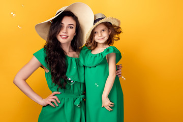 Beautiful mother standing with little girl in same green dresses and straw hats in studio with...