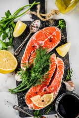 Raw salmon fish steaks with lemon, herbs, olive oil, ready for grill, slate cutting board, white marble background copy space above