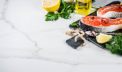 Raw salmon fish steaks with lemon, herbs, olive oil, ready for grill, slate cutting board, white marble background copy space above