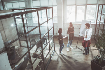 Business tasks. Full length top view of concentrated young colleagues are discussing in office. They are standing near flipchart while man is pointing on it. Women are listening him attentively