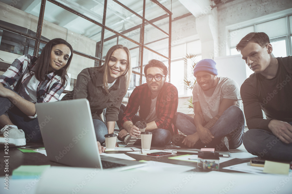 Poster low angle of optimistic young creative team are sitting on floor in office and looking at screen of 