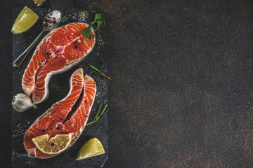 Raw salmon fish steaks with lemon, herbs, olive oil, ready for grill, slate cutting board, dark rusty background copy space