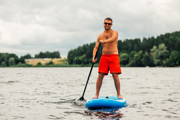 a man in shorts floats standing on a sapsurf on   lake in cloudy weather.