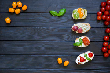 appetizer bruschettas with tomatoes, salmon, creamy cheese, basil, grapes, blue cheese, pear, avocado with fresh cherries tomatoes on black wooden background. Italian cuisine, top view, copy space.