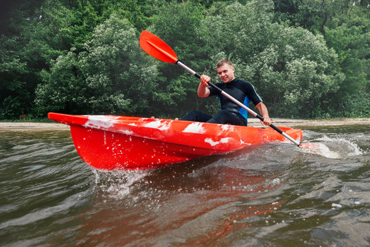 cheerful guy in   wetsuit sits in a red kayak and row with a paddle