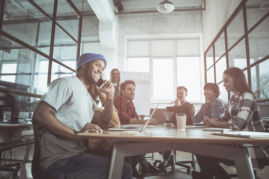 Pleasant Call. Low Angle Of Friendly Young Positive Team Is Sitting Around Table In Office And Discussing Business Project. Focus On Cheerful African Guy Is Talking On Modern Mobile Phone With Smile