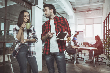 Look here. Cheerful man and woman are talking while standing in office. Female is drinking espresso while male is holding tablet and pointing on screen of gadget. Colleagues are sitting in background