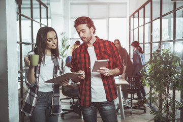 Two professional colleagues are standing together in office. Woman is drinking coffee while holding folder with document. Man is holding tablet and giving advices to his coworker about new project