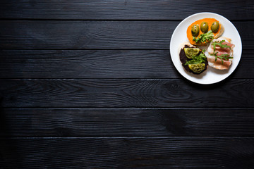 Plate with italian bruschettas with olive pasta, pesto, ham, parma, rucola, humus and rocked salad on black wooden background. Top view, copy space.