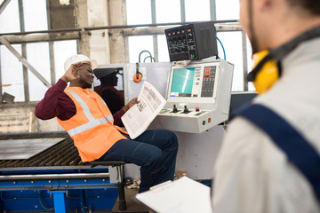 Positive carefree African-American automated machine operator waving hand with indifference while reading newspaper and talking to colleague at workplace