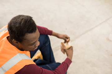 Obraz premium Directly above of pensive calm African-American construction worker in orange vest sitting on ground and eating from box during lunch break