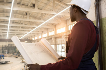 Rear view of pensive purposeful young African-American construction manager in hardhat holding blueprint with building plan and looking around at construction site